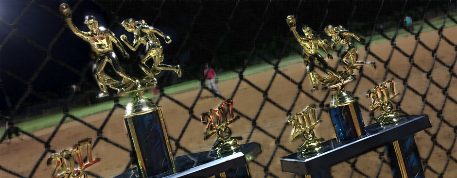 First and second place softball trophies with ballfield in background through fence.